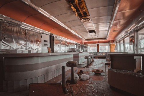 Abandoned diner interior with worn-out stools, debris on the floor, and a retro design featuring metallic walls and ceiling.