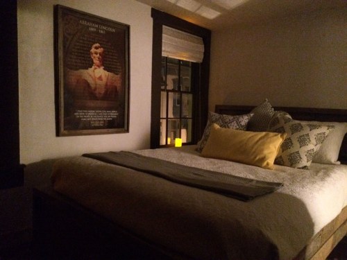 A cozy bedroom with a bed, decorative pillows, and a poster of Abraham Lincoln on the wall, softly lit by a candle.