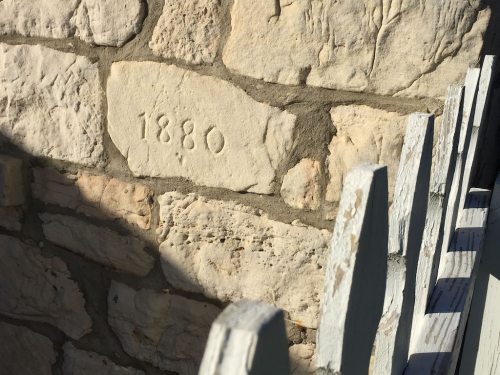 Close-up of a stone wall with the year "1880" engraved, alongside a wooden picket fence.