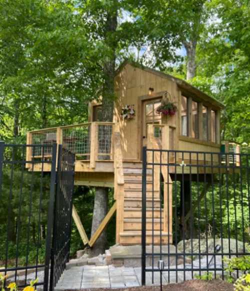 A wooden treehouse with large windows, surrounded by trees, accessed by a staircase and enclosed by a black fence.