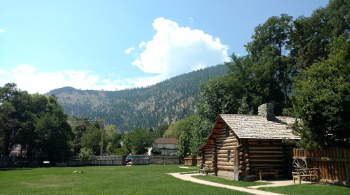 A rustic log cabin surrounded by green grass and trees, with mountains in the background under a blue sky.