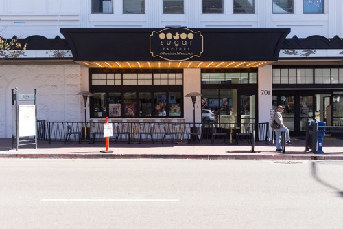 Exterior of Sugar Factory restaurant with outdoor seating, bright signage, and a person sitting nearby on the sidewalk.