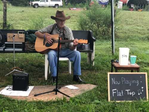 A man in a cowboy hat plays guitar on a bench, with a sign reading "Now Playing: Flat-Top" nearby.