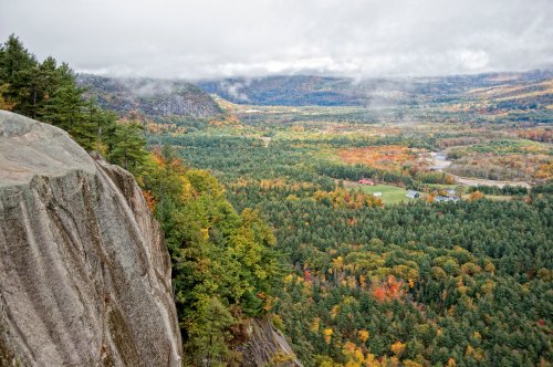 A panoramic view of a lush valley with autumn foliage, mountains, and a river under a cloudy sky.