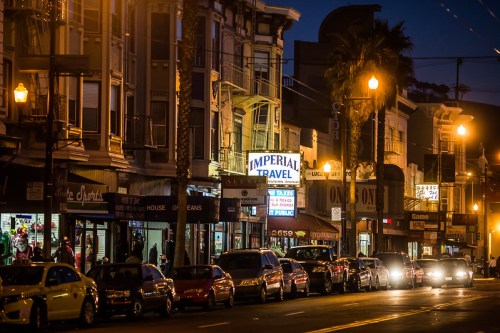 A street scene at night featuring shops, palm trees, and parked cars, with "Imperial Travel" illuminated in the background.