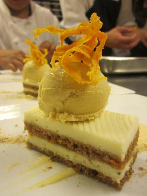 A layered dessert with cream, nuts, and a scoop of ice cream topped with orange peel curls on a white plate.