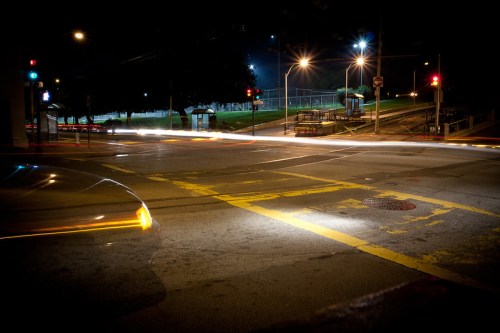 Nighttime street scene with blurred car lights, traffic signals, and an empty bus stop in a quiet urban area.
