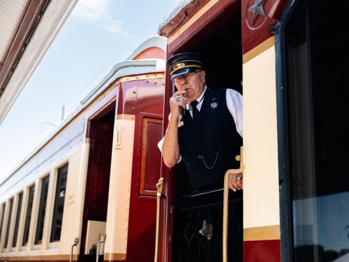 A train conductor stands at the door of a vintage train, signaling with a finger to his lips.