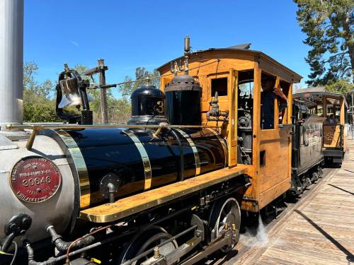A vintage steam locomotive with a wooden cab, parked on a sunny day, surrounded by trees and power lines.