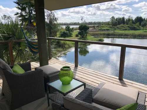 A cozy balcony with wicker furniture overlooks a serene lake surrounded by greenery and blue skies.