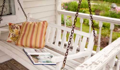 A white swing bench with a colorful pillow and an open book, surrounded by a lush garden.