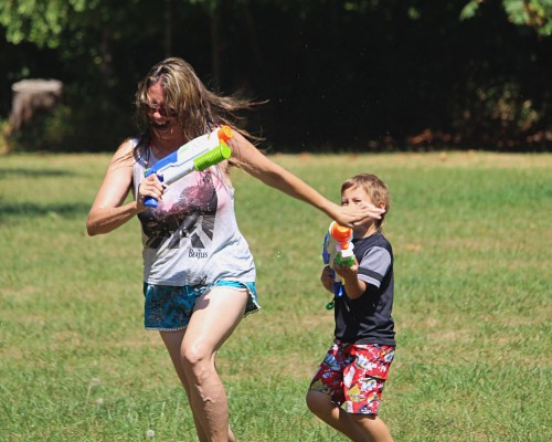 A woman and a boy playfully chase each other with water guns in a grassy field on a sunny day.