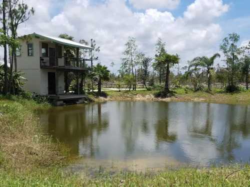 A small house by a pond, surrounded by trees and grass under a partly cloudy sky.