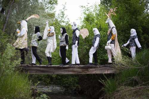 A group of people in elaborate costumes walking in a line on a wooden bridge surrounded by greenery.