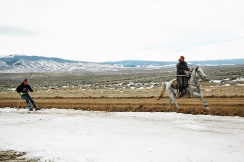 A person on horseback races alongside another person on skis in a snowy, mountainous landscape.