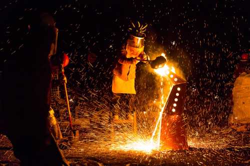 A worker in protective gear pours molten metal, creating sparks against a dark background.