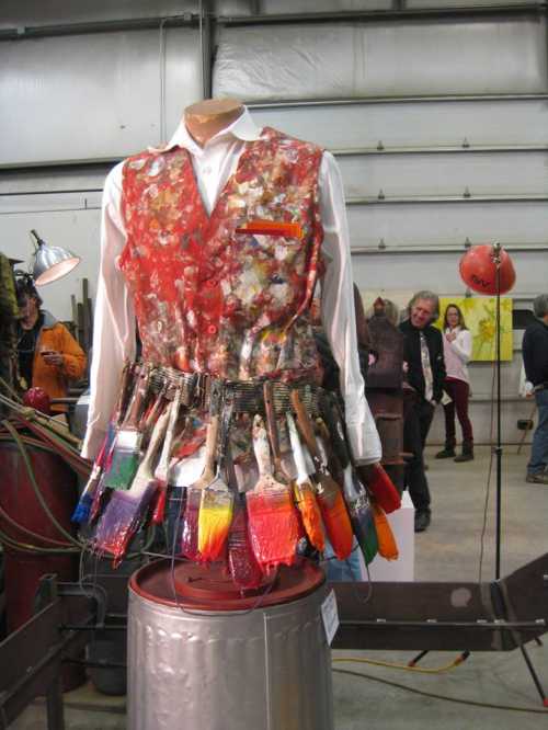 A colorful artist's vest adorned with paintbrushes, displayed on a mannequin in an art exhibition.