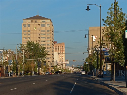 A wide street lined with trees and buildings, featuring a mix of modern and older architecture under a clear blue sky.