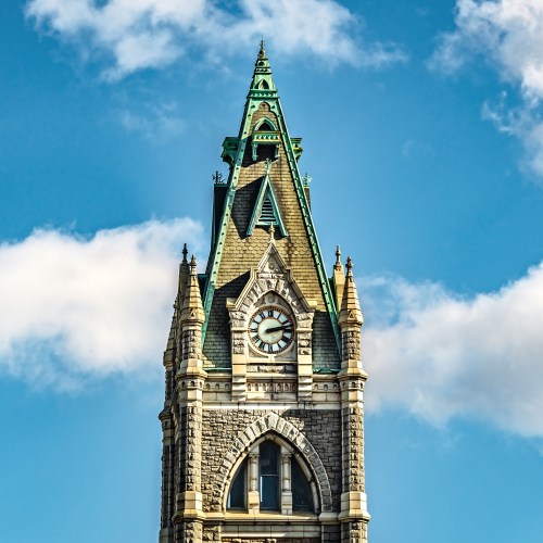 A tall clock tower with a green spire, featuring intricate stonework and a large clock face against a blue sky.