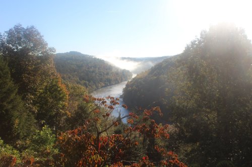 A serene river winds through misty hills, framed by trees with autumn foliage under a bright sky.