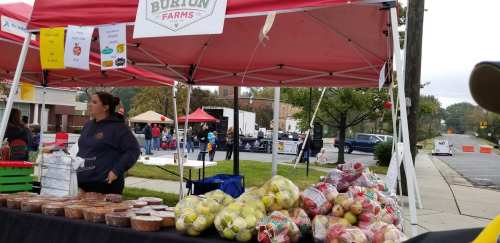 A farmer's market stall with fresh produce, including apples and baked goods, under a red tent. People are in the background.