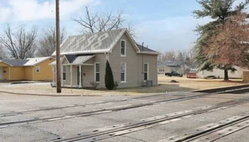 A small, light-colored house at a street corner near railroad tracks, with a few trees and other buildings in the background.