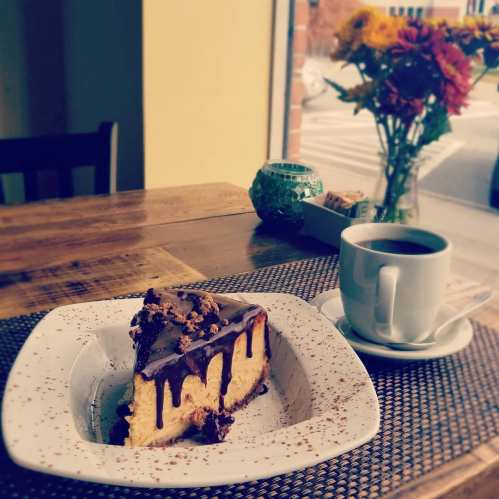 A slice of chocolate-drizzled cake on a plate, with a cup of coffee and a vase of flowers in the background.