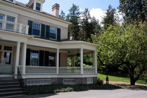 A historic house with a porch, surrounded by trees and greenery on a sunny day.