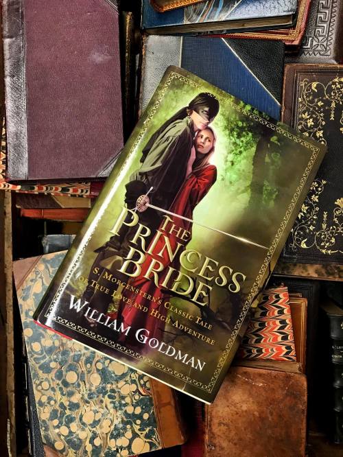 A copy of "The Princess Bride" by William Goldman rests on a stack of vintage books with ornate covers.