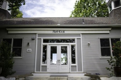 Front view of a building labeled "Playhouse," featuring large glass doors and a gray exterior surrounded by trees.