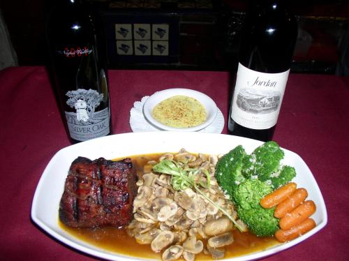 A gourmet meal featuring grilled steak, mushrooms, broccoli, carrots, and a side dish, with two bottles of wine in the background.