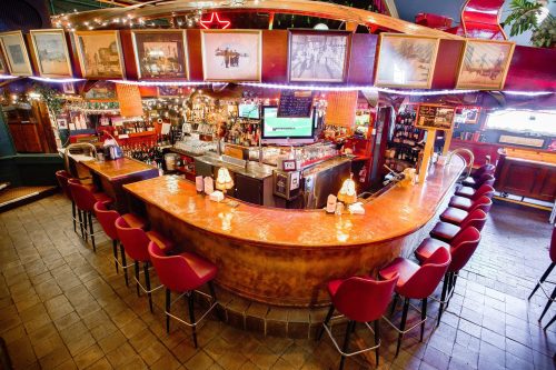 A cozy bar with a curved counter, red stools, and framed photos, illuminated by soft lights in a warm atmosphere.
