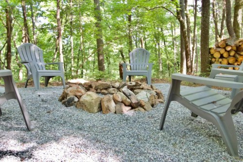 A cozy fire pit surrounded by gravel and four gray chairs in a wooded area.