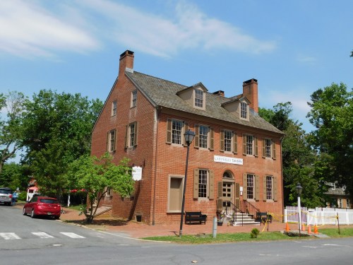 Historic brick building with a gabled roof, surrounded by trees and parked cars, under a clear blue sky.