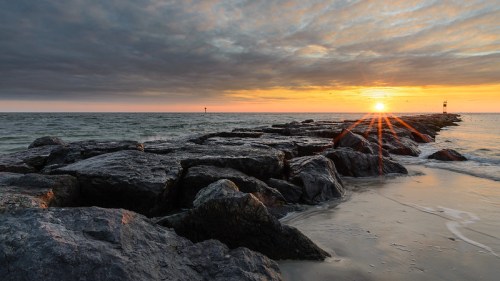 A rocky jetty extends into the ocean at sunset, with vibrant colors in the sky and gentle waves lapping at the shore.