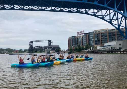 A group of people kayaking in colorful kayaks under a blue bridge, with buildings in the background.