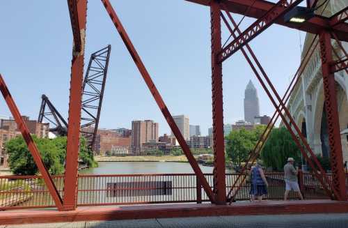 View of a river and city skyline framed by a red metal bridge structure, with two people walking along the path.