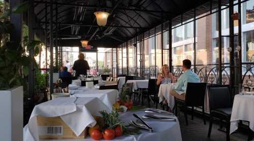 A cozy restaurant patio with tables set for dining, featuring plants and a couple enjoying a meal in the background.