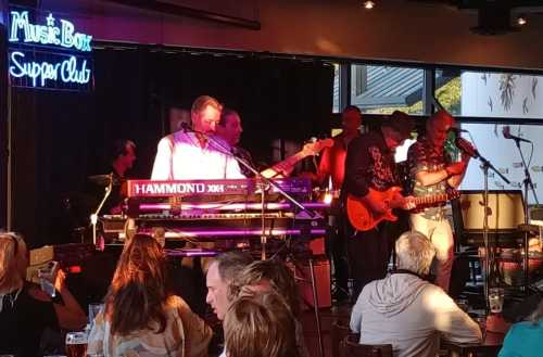 A live band performs on stage at a venue, with a neon "Music Box Super Club" sign in the background. Audience enjoying the show.