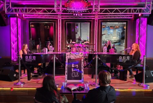 Two women perform at pianos on stage, with a drummer in the background, in a vibrant, illuminated venue.