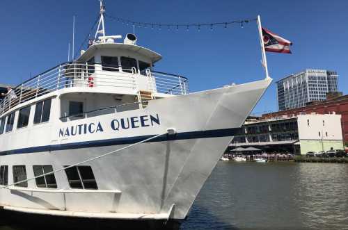 A white boat named "Nautica Queen" docked by a waterfront with a clear blue sky and buildings in the background.