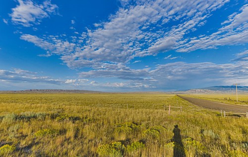 A vast landscape with grassy fields, distant mountains, and a blue sky filled with clouds, featuring a shadow in the foreground.