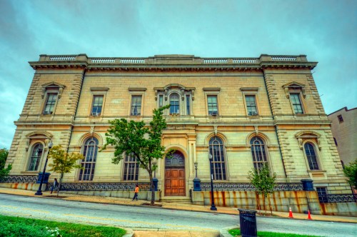 Historic stone building with large windows and a wooden door, set against a cloudy sky. Trees line the sidewalk.