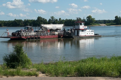 A barge and tugboat navigate a calm river under a blue sky with scattered clouds, surrounded by greenery.