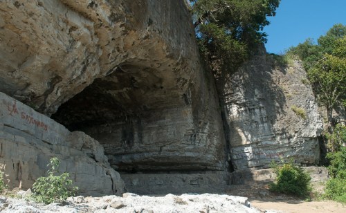 A rocky cliffside with overhanging ledges and greenery, under a clear blue sky.