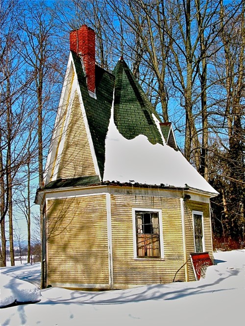 A small, snow-covered house with a steep roof and chimney, surrounded by bare trees in a winter landscape.