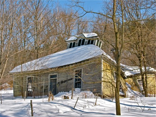 An old, weathered house with a slanted roof, partially covered in snow, surrounded by bare trees in a winter landscape.