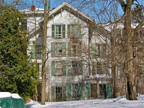 An old, abandoned building with green shutters, surrounded by snow and bare trees under a clear blue sky.