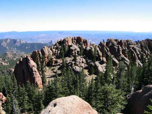 A panoramic view of rugged rock formations surrounded by dense evergreen trees and distant mountains under a clear blue sky.