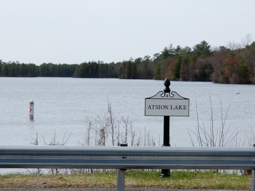 A sign for Atsion Lake beside a calm body of water, with trees lining the shore in the background.